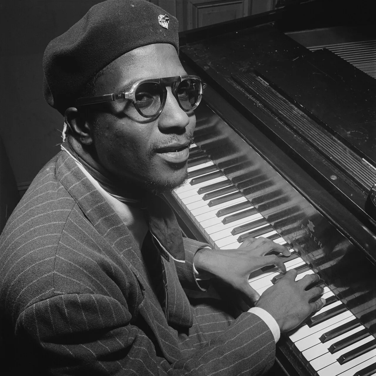 Thelonious Monk seated at the piano, in a beret and glasses, looking at the camera